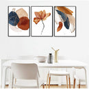 Set of 3 abstract photo frames with brown, navy, and beige tones hanging above a white dining table with chairs.