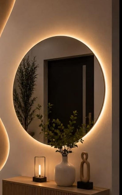 A modern round wall mirror with a warm glowing LED light border, mounted on a beige wall above a wooden console table decorated with a white vase, a small candle holder, and a decorative sculpture.
