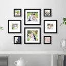 A set of 8 black picture frames of varying sizes arranged in a collage pattern on a white wall above a white shelf with books, a teapot, and decorative items.