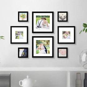 A set of 8 black picture frames of varying sizes arranged in a collage pattern on a white wall above a white shelf with books, a teapot, and decorative items.