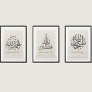 Three framed calligraphy artworks with Arabic script, displayed on a wall. Each frame has a different color: black, white, and gold.